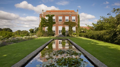 Georgian-style red brick house covered in climbing ivy, viewed from a formal garden with a long rectangular reflecting pool filled with lily pads in the foreground, mirrored in the still water, under a bright blue sky with scattered clouds.